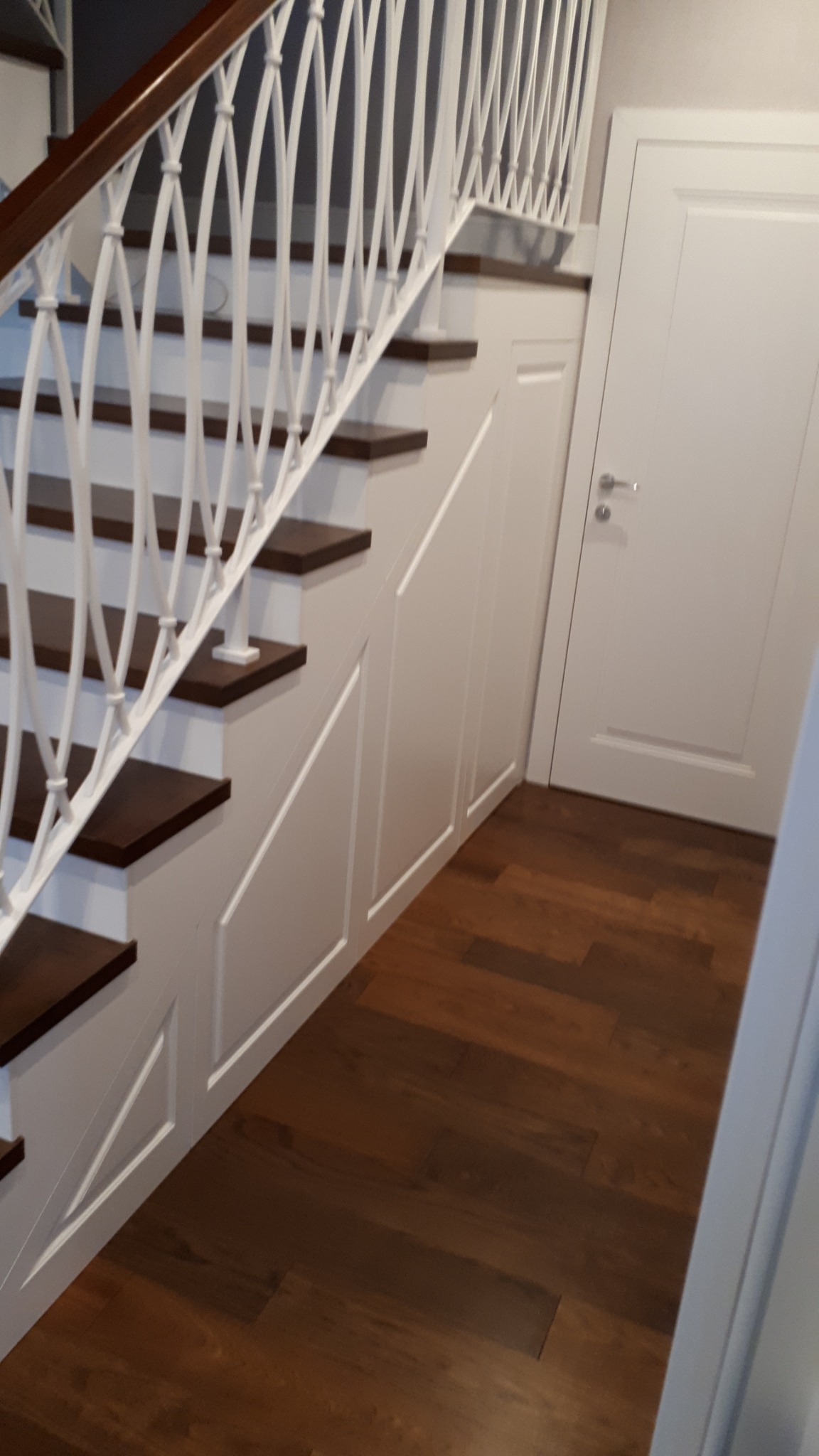 Klasyczne schody z ciemnymi stopniami i białą balustradą z dekoracyjnymi elementami, zabudowane szafki pod schodami z białymi frontami, parkiet z ciemnego drewna.