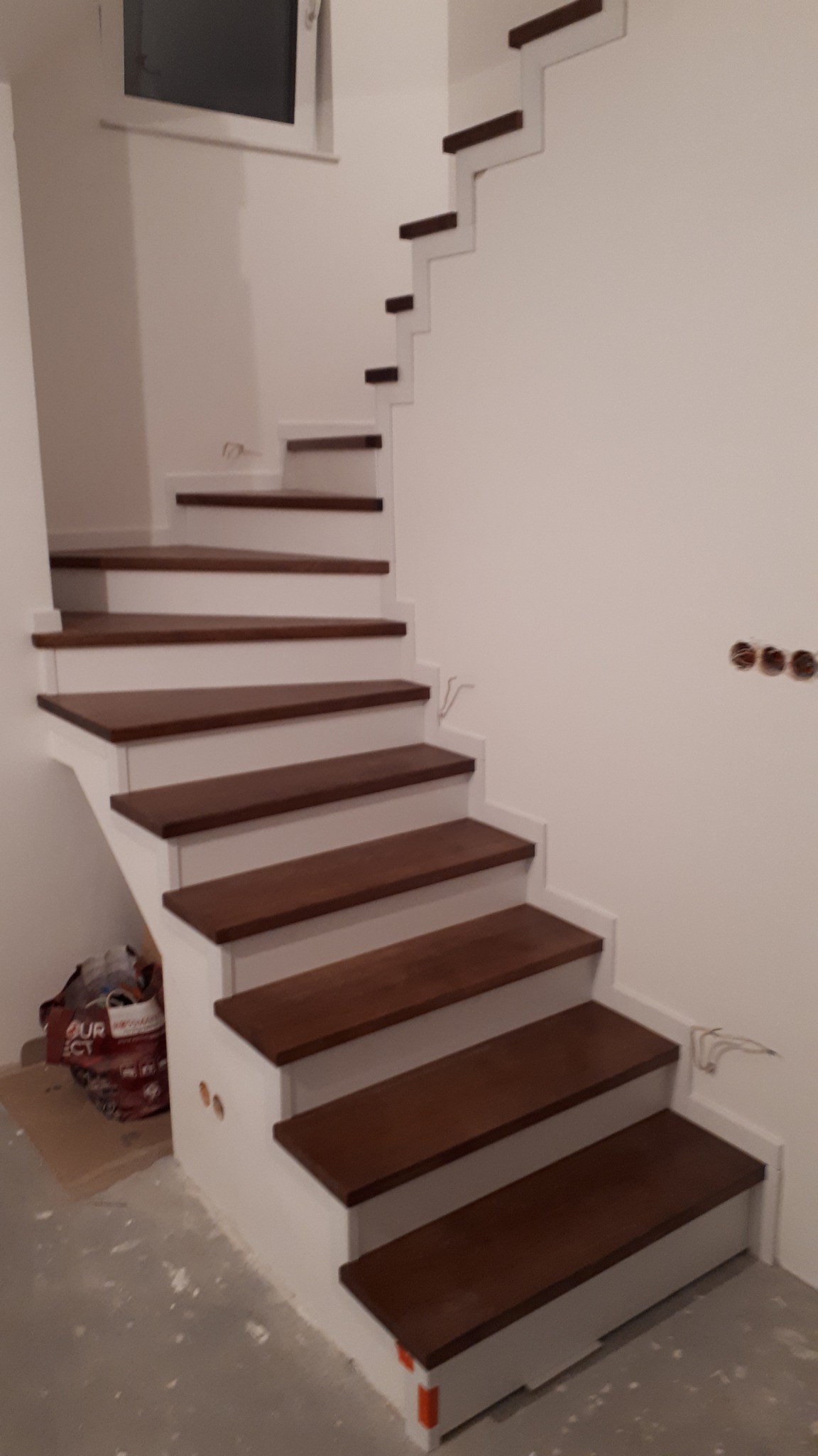 Nowoczesne schody o konstrukcji wspornikowej z ciemnego drewna i białych elementów, widoczne niedokończone wykończenie ściany i podłogi.