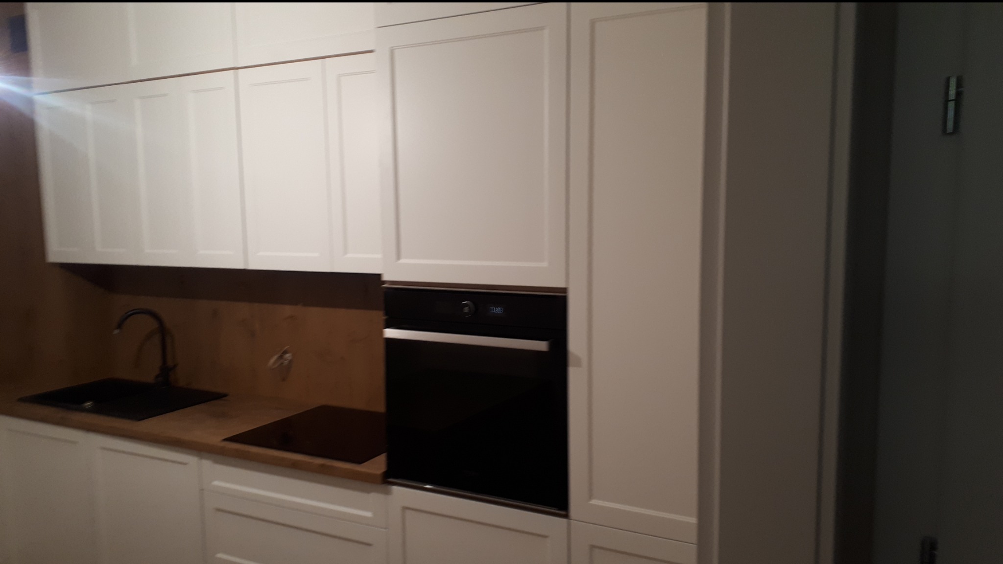 Nowoczesna kuchnia z białymi szafkami, czarnym zlewem i płytą indukcyjną, oraz zabudowanym piekarnikiem. Uwagę zwraca drewniany panel nad blatem.