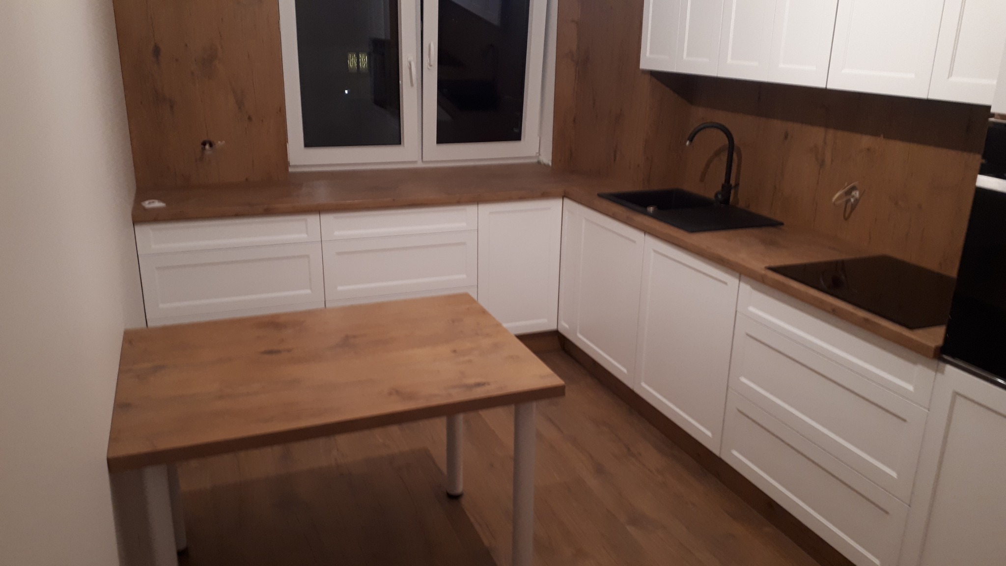 Nowoczesna kuchnia z białymi szafkami i blatem imitującym drewno, czarny zlew i płyta indukcyjna, stół z blatem w kolorze blatu kuchennego, widok na okno z zabudową.