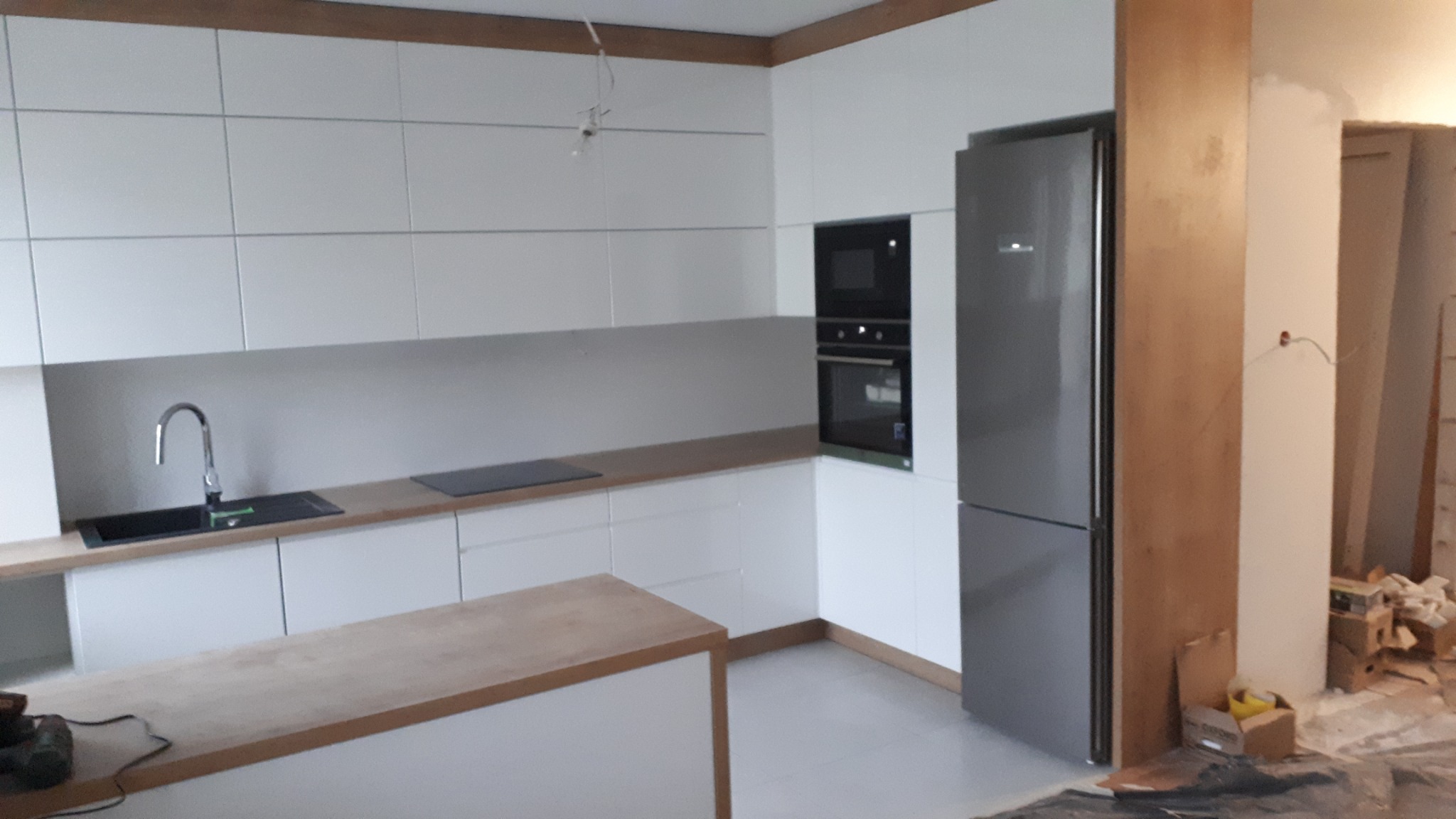 Nowoczesna kuchnia w trakcie montażu: białe szafki z drewnianymi blatami, wbudowany sprzęt AGD i wyspa kuchenna, widoczne elementy wykończeniowe i narzędzia.