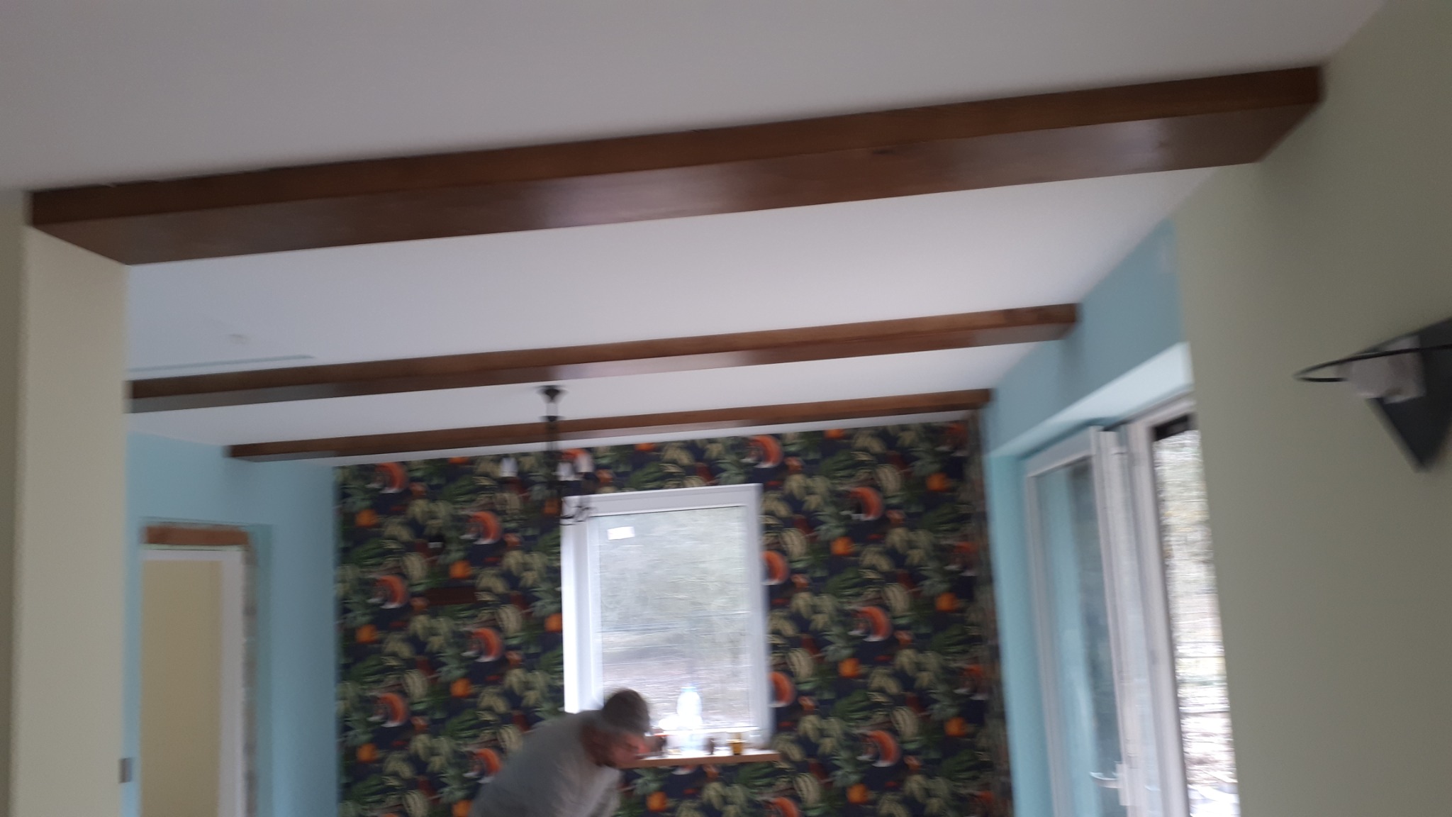 Wnętrze pokoju z zamontowanymi dekoracyjnymi belkami stropowymi z drewna, widoczny fragment osoby pracującej przy oknie