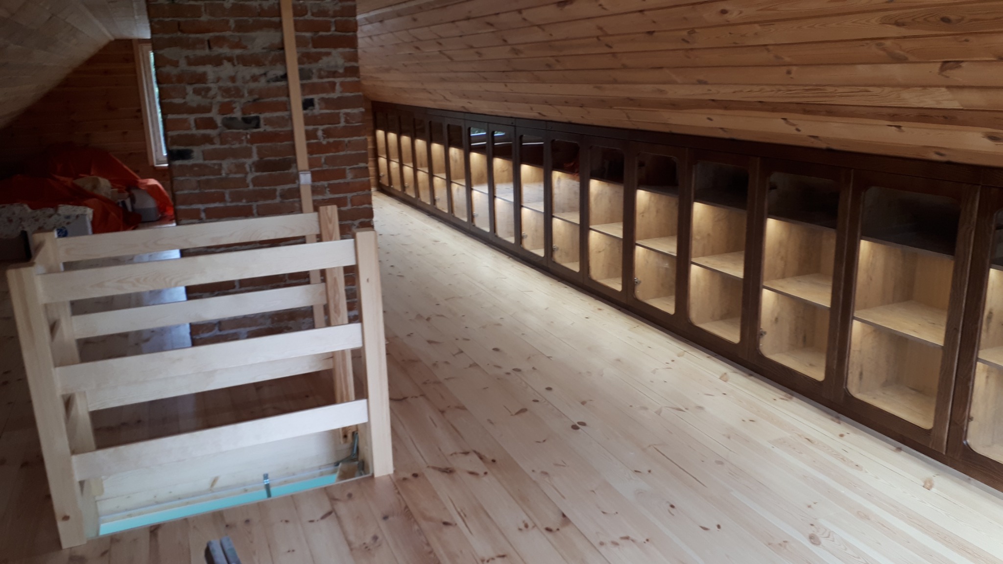 Poddasze wykończone drewnem: boazeria, podłoga z jasnych desek, zabudowa szafek z oświetleniem oraz fragment surowej ściany z cegły i drewniana konstrukcja przypominająca barierkę.