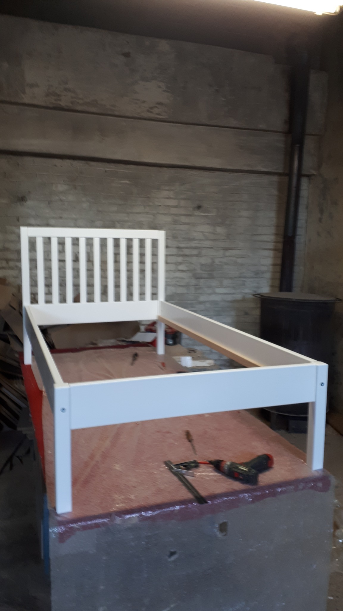 Biała rama łóżka z zagłówkiem w trakcie montażu, widoczne narzędzia i surowe otoczenie warsztatowe.