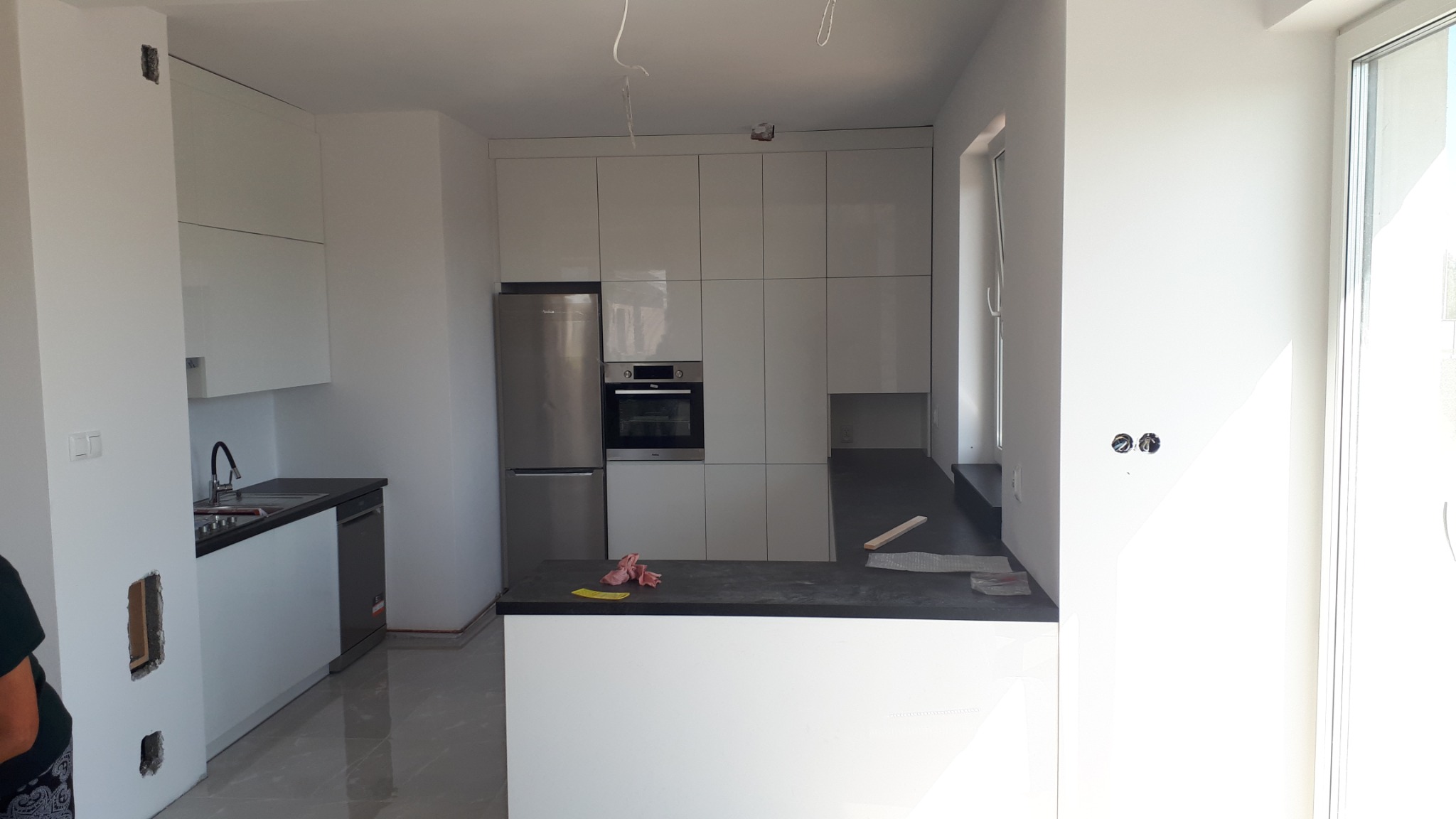 Nowoczesna, jasna kuchnia z białymi szafkami, czarnym blatem i srebrnymi urządzeniami w trakcie montażu, widoczne wystające kable i niedokończone ściany.