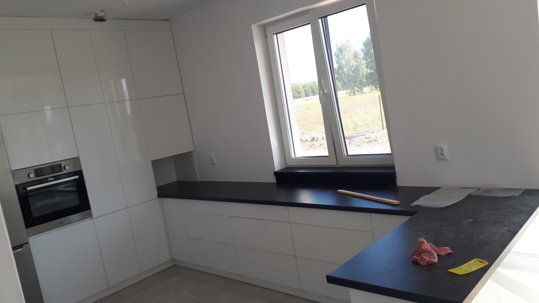 Nowoczesna kuchnia z białymi, lakierowanymi frontami szafek, czarnym blatem i wbudowanym piekarnikiem; widok na okno z widokiem na zewnątrz, na blacie leżą ściereczka i miarka.