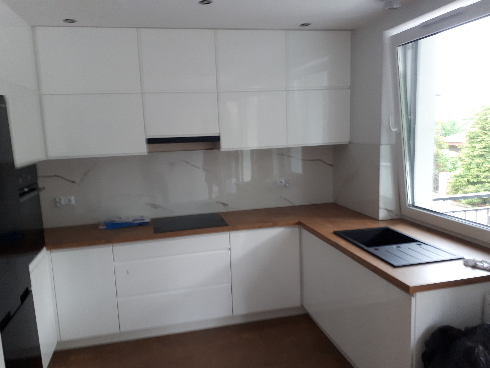 Nowoczesna, biała kuchnia z drewnianym blatem, minimalistyczny design, widok na zewnątrz przez okno.