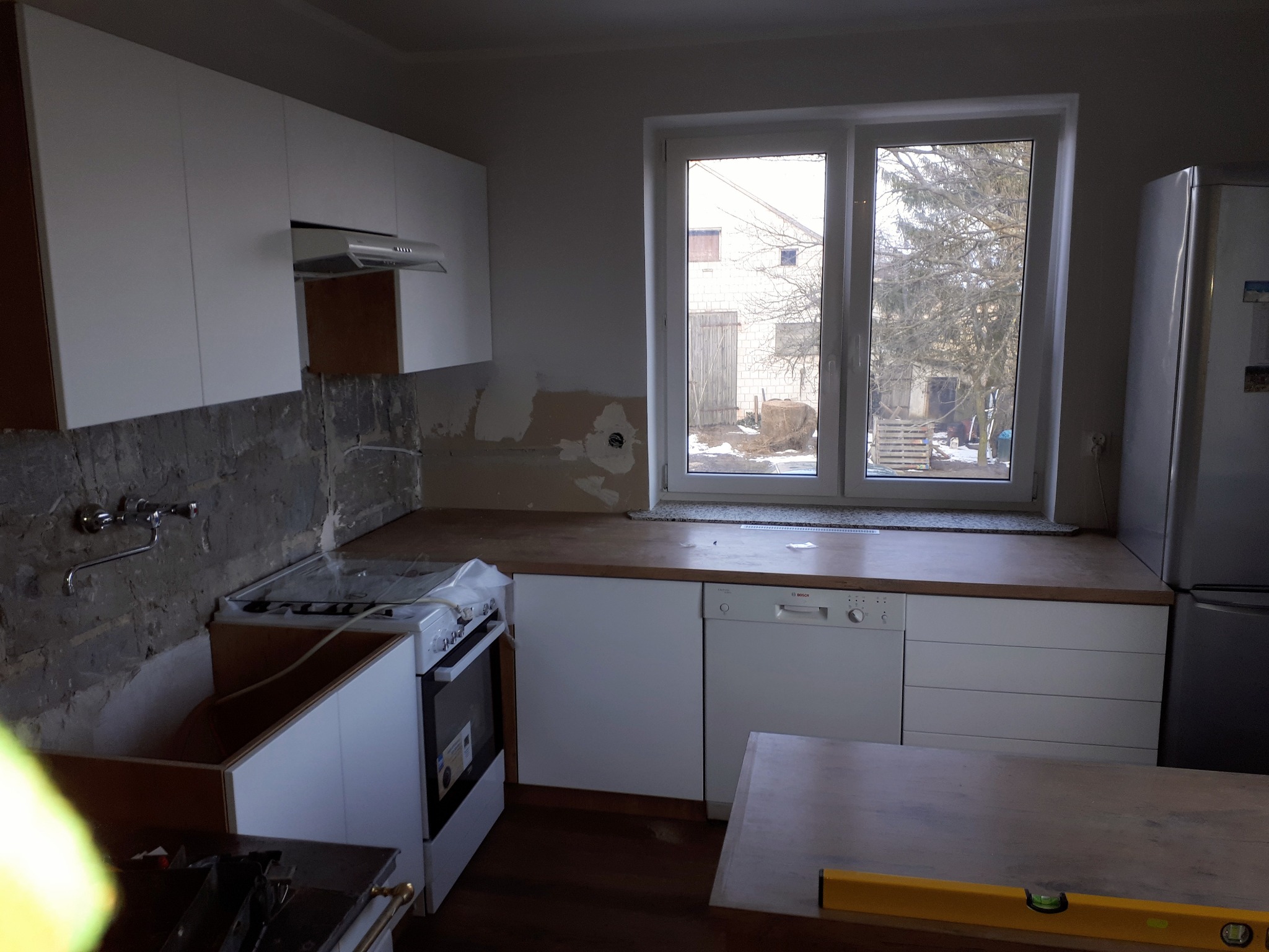 Wnętrze kuchni w trakcie remontu: białe szafki kuchenne, częściowo wykończone ściany, widoczne instalacje, okno z widokiem na budynek gospodarczy i zimowy krajobraz, poziomica na blacie.
