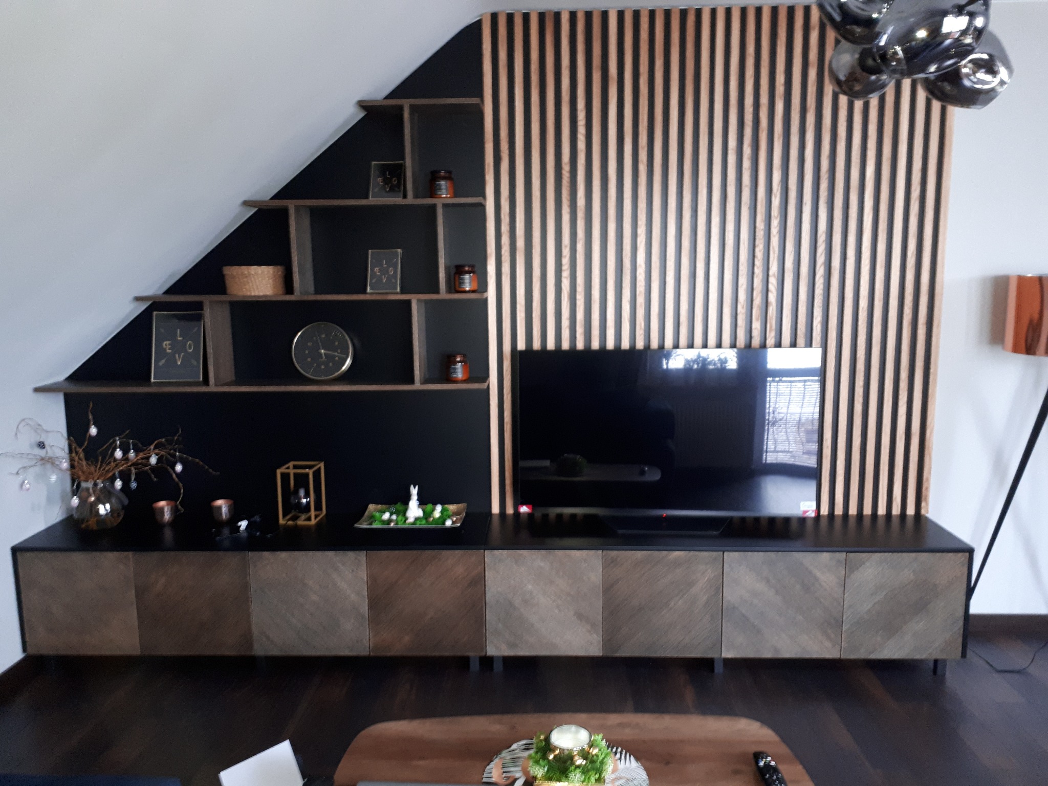 Nowoczesny salon z drewnianą ścianą z lameli, półkami pod skosem i szafką RTV z frontami w jodełkę.