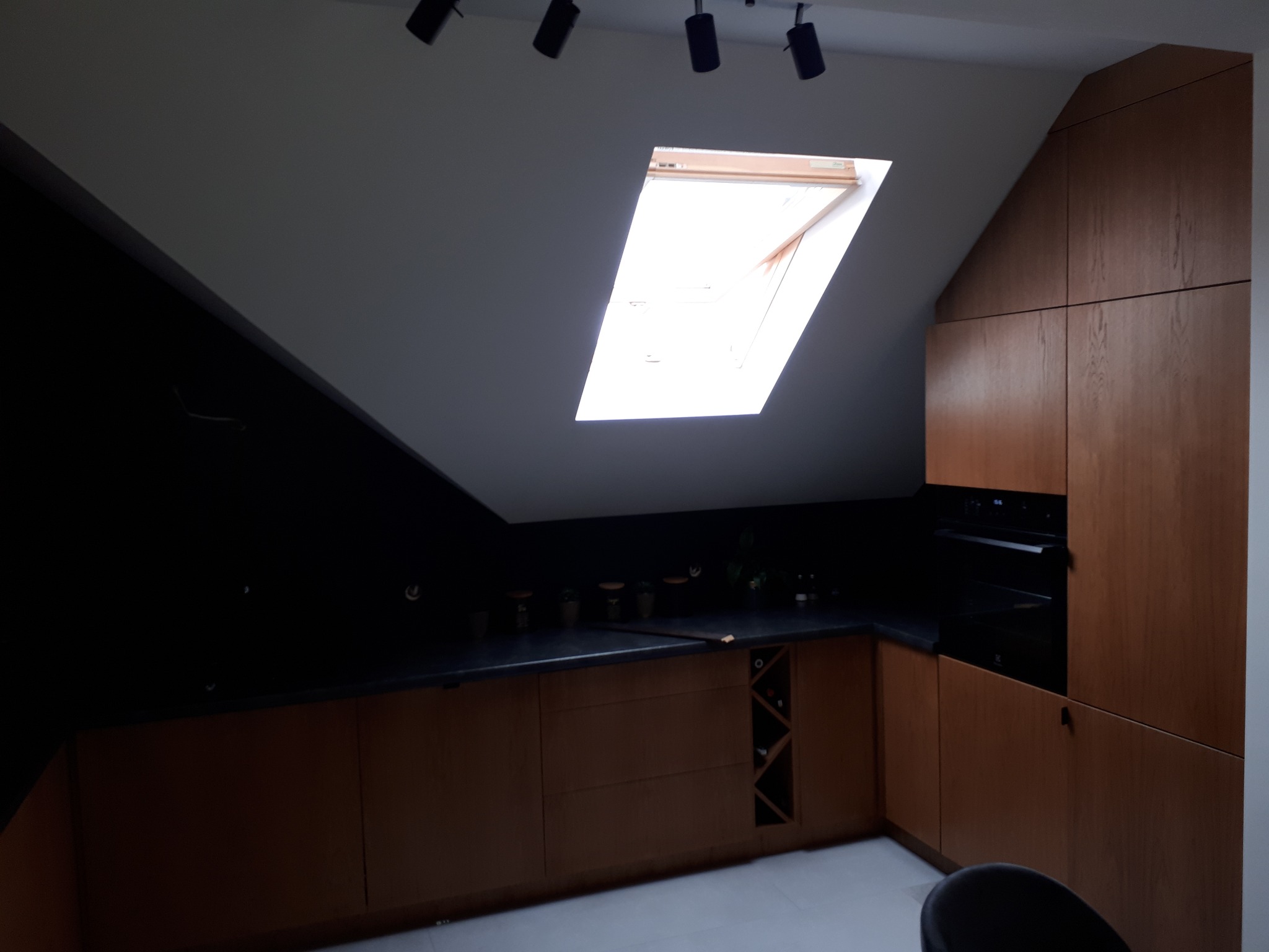 Kuchnia w skosach z drewnianymi frontami, czarnym blatem i oknem dachowym, oświetlona reflektorami punktowymi.