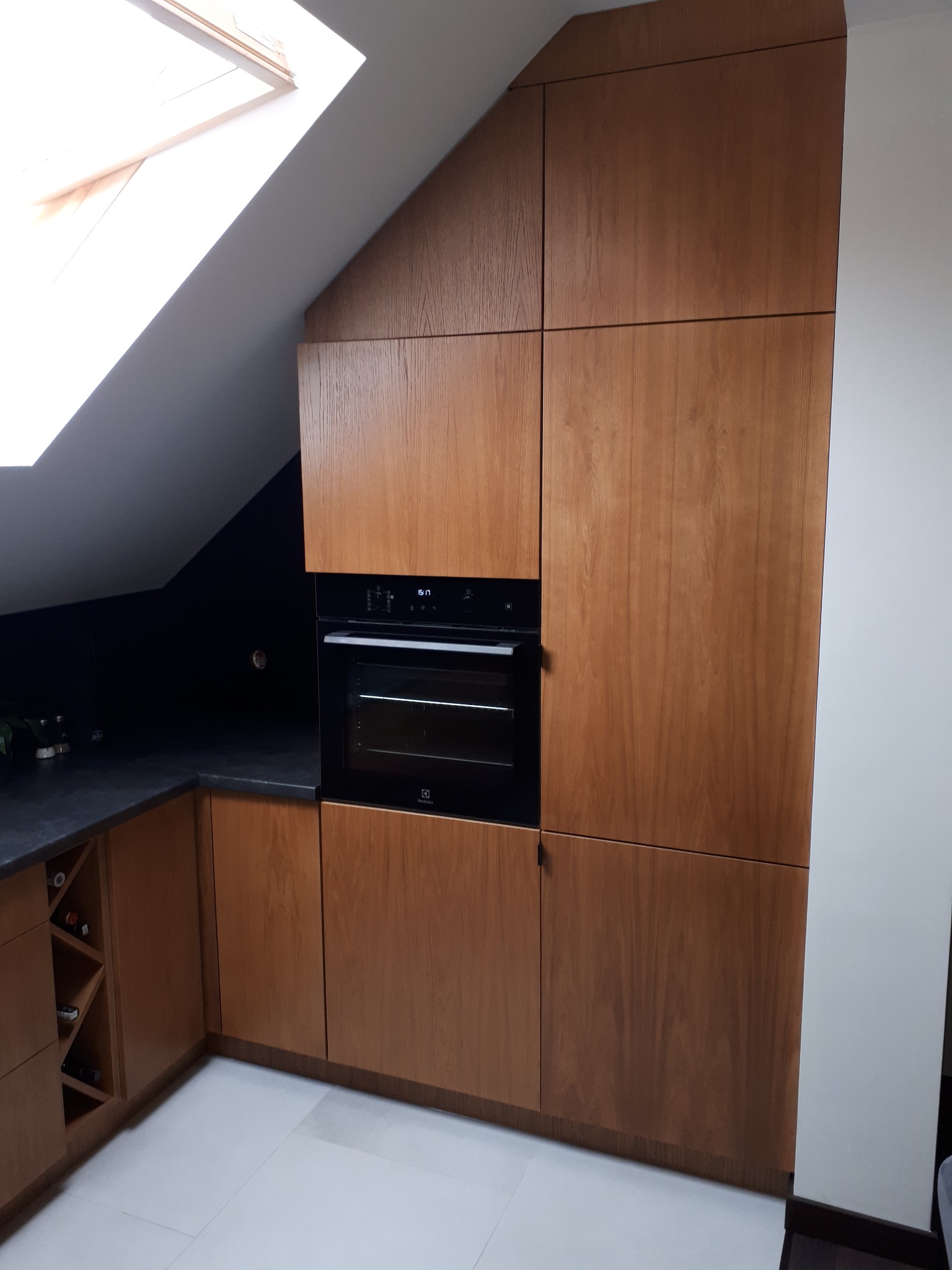Drewniana zabudowa kuchenna na poddaszu z piekarnikiem Electrolux, ciemnym blatem i półkami na wino.