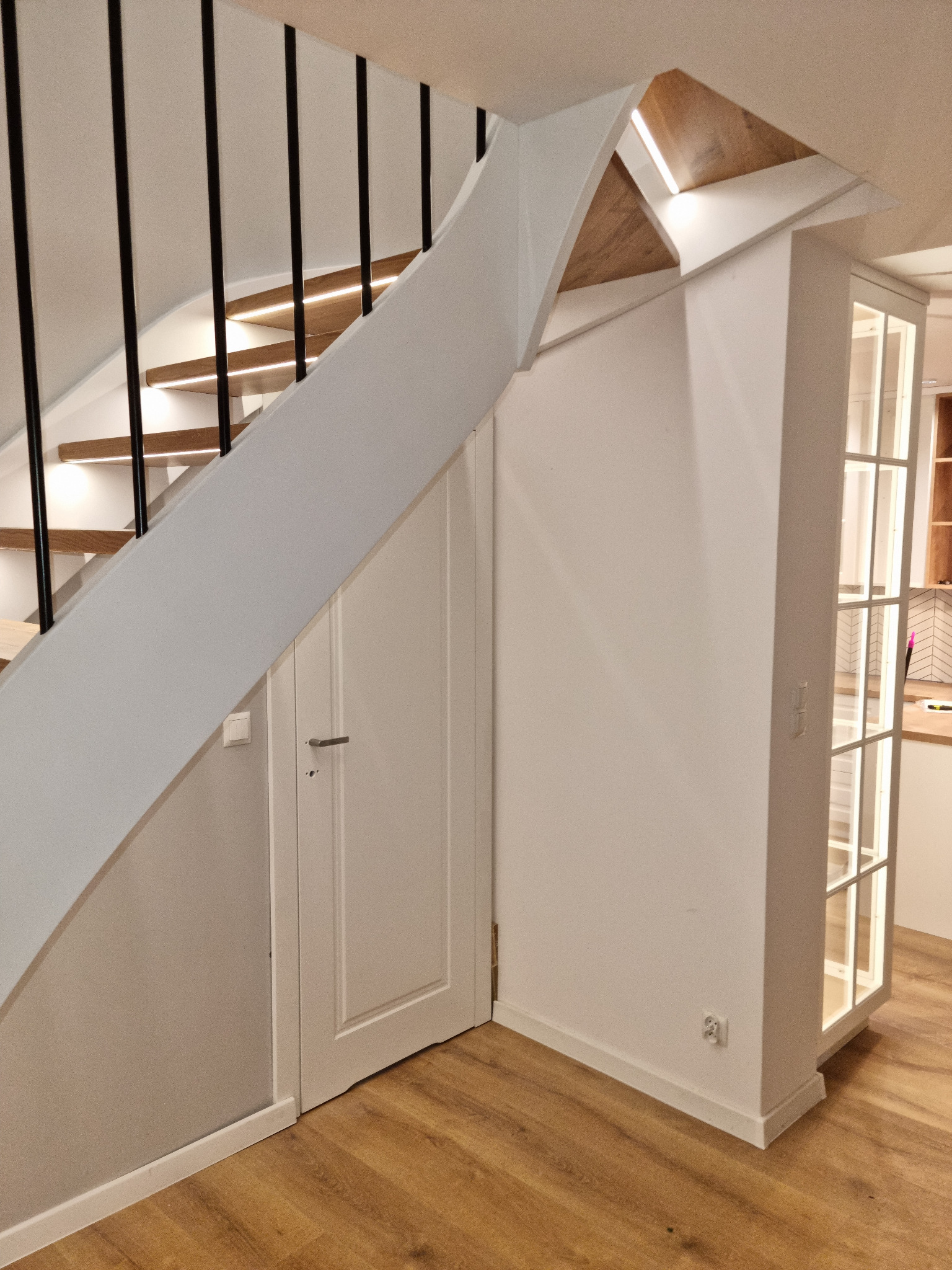 Nowoczesne schody z drewnianymi stopniami i czarną balustradą, oświetlone od spodu. Pod schodami białe drzwi i zabudowa z przeszklonymi drzwiami. Jasne wnętrze.