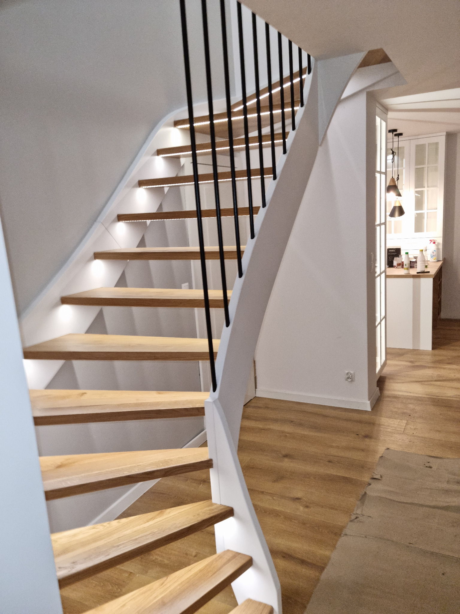 Nowoczesne schody drewniane z podświetleniem LED, czarne balustrady, białe boki, widok na kuchnię w tle. Jasne drewno stopni, minimalistyczny design.