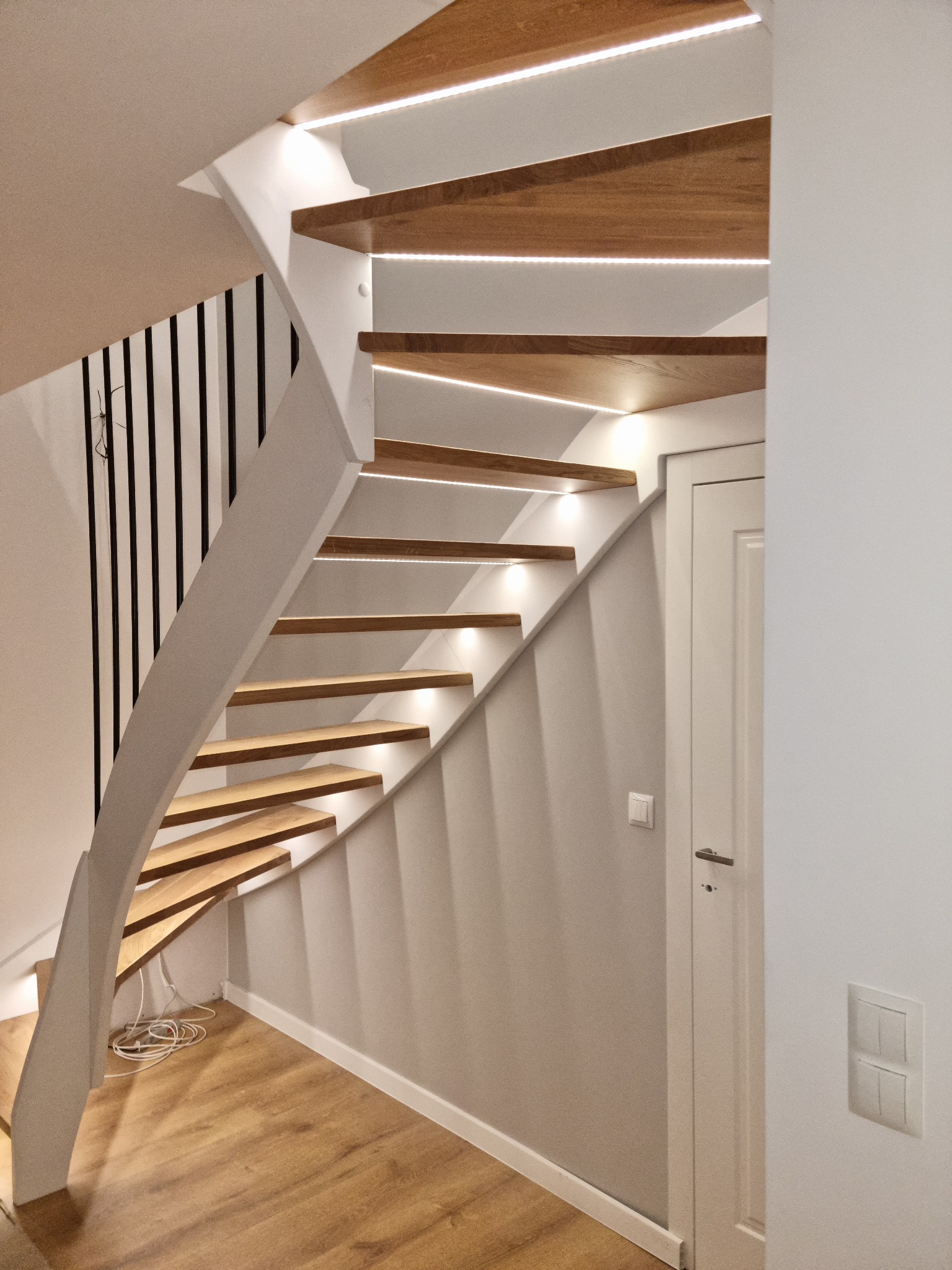 Nowoczesne schody drewniane z białymi elementami i subtelnym oświetleniem LED, widoczne z dołu, z drewnianą podłogą i białymi ścianami w tle.