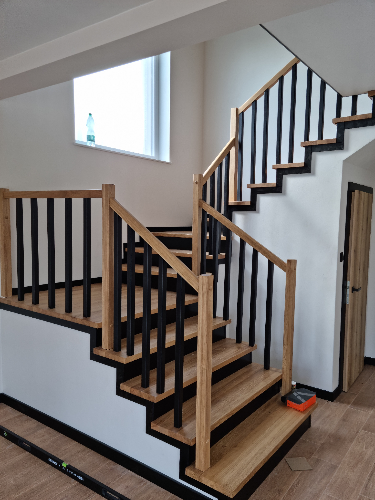 Nowoczesne schody z drewnianymi stopniami i czarnymi balustradami, oświetlone naturalnym światłem z okna. Widoczny poziomica i narzędzia, co sugeruje etap końcowy montażu.