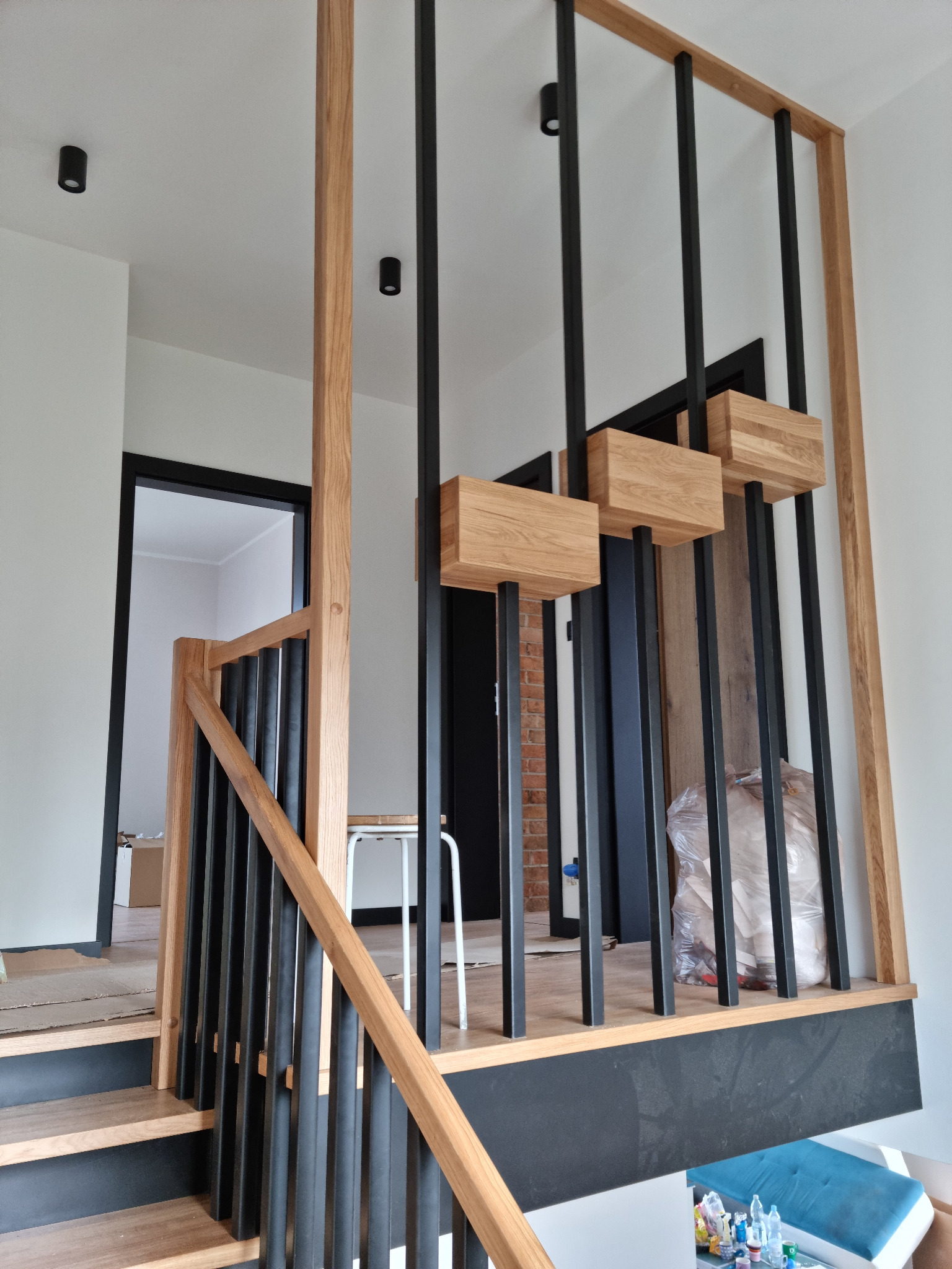 Nowoczesne schody z drewnianymi stopniami i czarnymi, metalowymi balustradami. Elementy drewniane z wyraźnym usłojeniem. Widoczne oświetlenie sufitowe.