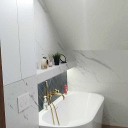 Studio Projektowe Decorfun - Biała wanna wolnostojąca z złotą armaturą w nowoczesnej łazience na poddaszu, wykończonej płytkami imitującymi marmur. Widoczna toaleta podwieszana i półka z dekoracjami.