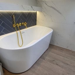 Studio Projektowe Decorfun - Nowoczesna łazienka z białą wanną wolnostojącą, złotą armaturą i marmurowymi płytkami. Ciemne płytki ułożone w jodełkę nad wanną, podświetlone listwą LED.