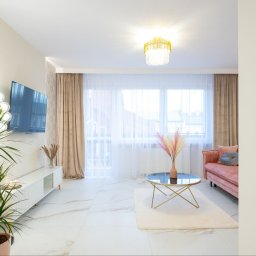 Studio Projektowe Decorfun - Jasne wnętrze salonu z marmurową podłogą, różową sofą i złotymi akcentami: lampa, stolik kawowy. Duże okno z zasłonami w odcieniach beżu. Roślina doniczkowa po lewej.