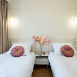 Studio Projektowe Decorfun - Jasna sypialnia z dwoma łóżkami pojedynczymi, beżowe ściany z dekoracyjną listwą, okrągłe poduszki w kolorze brudnego różu, kwiaty w wazonie na szafce nocnej.