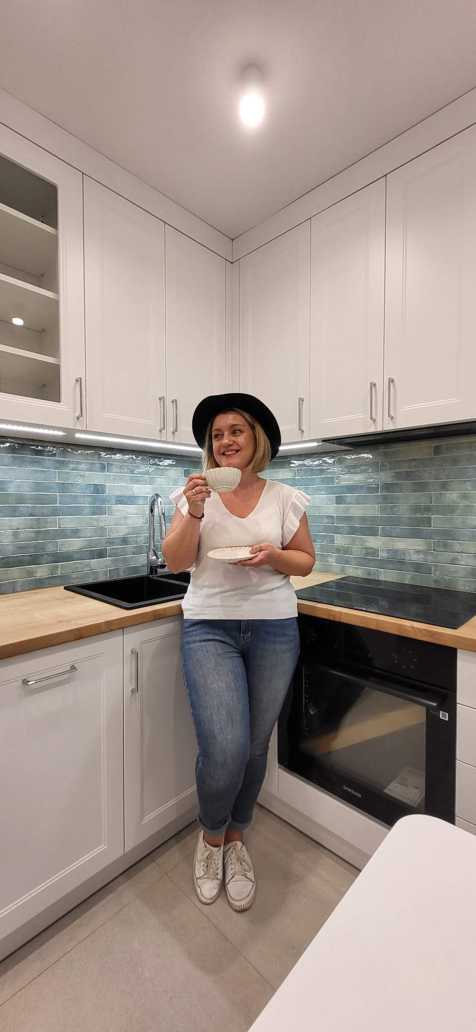 Jasna kuchnia z błękitnymi płytkami: kobieta w kapeluszu pije kawę, opierając się o blat. Minimalistyczny design z drewnianymi akcentami i subtelnym oświetleniem.