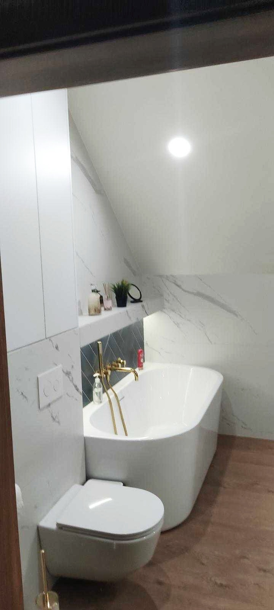 Biała wanna wolnostojąca z złotą armaturą w nowoczesnej łazience na poddaszu, wykończonej płytkami imitującymi marmur. Widoczna toaleta podwieszana i półka z dekoracjami.