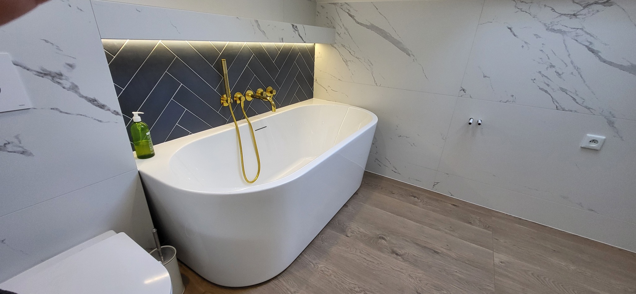 Nowoczesna łazienka z białą wanną wolnostojącą, złotą armaturą i marmurowymi płytkami. Ciemne płytki ułożone w jodełkę nad wanną, podświetlone listwą LED.