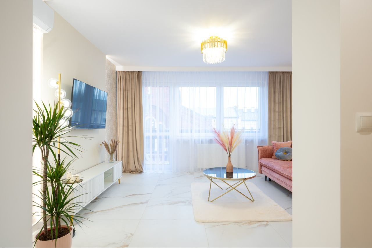 Jasne wnętrze salonu z marmurową podłogą, różową sofą i złotymi akcentami: lampa, stolik kawowy. Duże okno z zasłonami w odcieniach beżu. Roślina doniczkowa po lewej.