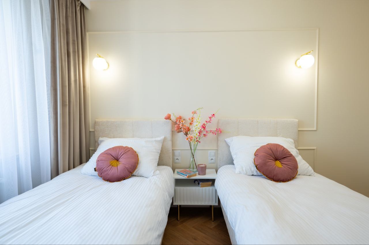 Jasna sypialnia z dwoma łóżkami pojedynczymi, beżowe ściany z dekoracyjną listwą, okrągłe poduszki w kolorze brudnego różu, kwiaty w wazonie na szafce nocnej.