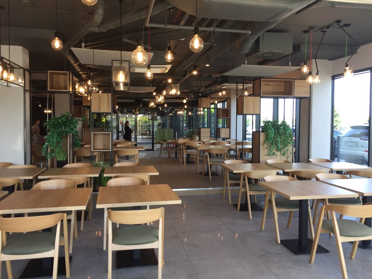 Jasne, przestronne wnętrze kawiarni z drewnianymi stolikami i krzesłami. Industrialny sufit z wiszącymi lampami. Dekoracyjne regały z roślinami. Duże okna wpuszczają naturalne światło.