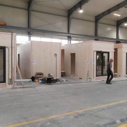 CASEHO INVEST - Wnętrze hali produkcyjnej z kilkoma surowymi, drewnianymi modułami domów w trakcie montażu, widoczny pracownik przechodzący obok.