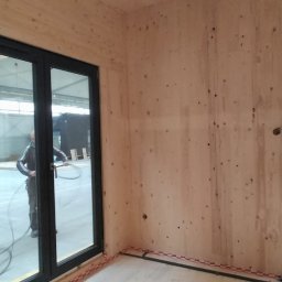 CASEHO INVEST - Wnętrze w surowym stanie z drewnianymi ścianami i czarnymi drzwiami balkonowymi; widoczny pracownik z wężem instalacyjnym w tle hali produkcyjnej.