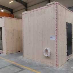 CASEHO INVEST - Wnętrze hali produkcyjnej z trzema surowymi modułami domów w trakcie montażu, widoczne okna, drzwi, instalacje elektryczne oraz oznaczenia na ścianach.