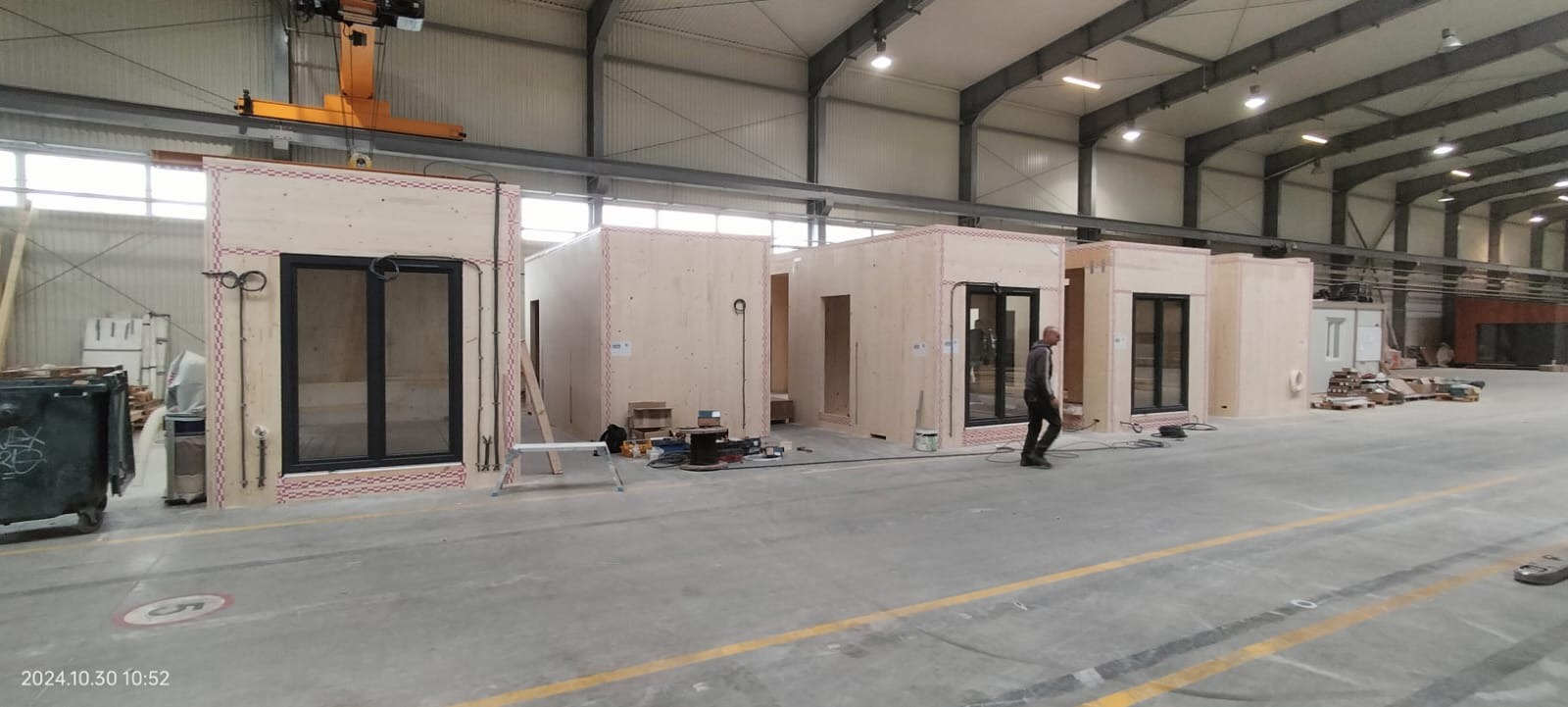 Wnętrze hali produkcyjnej z kilkoma surowymi, drewnianymi modułami domów w trakcie montażu, widoczny pracownik przechodzący obok.