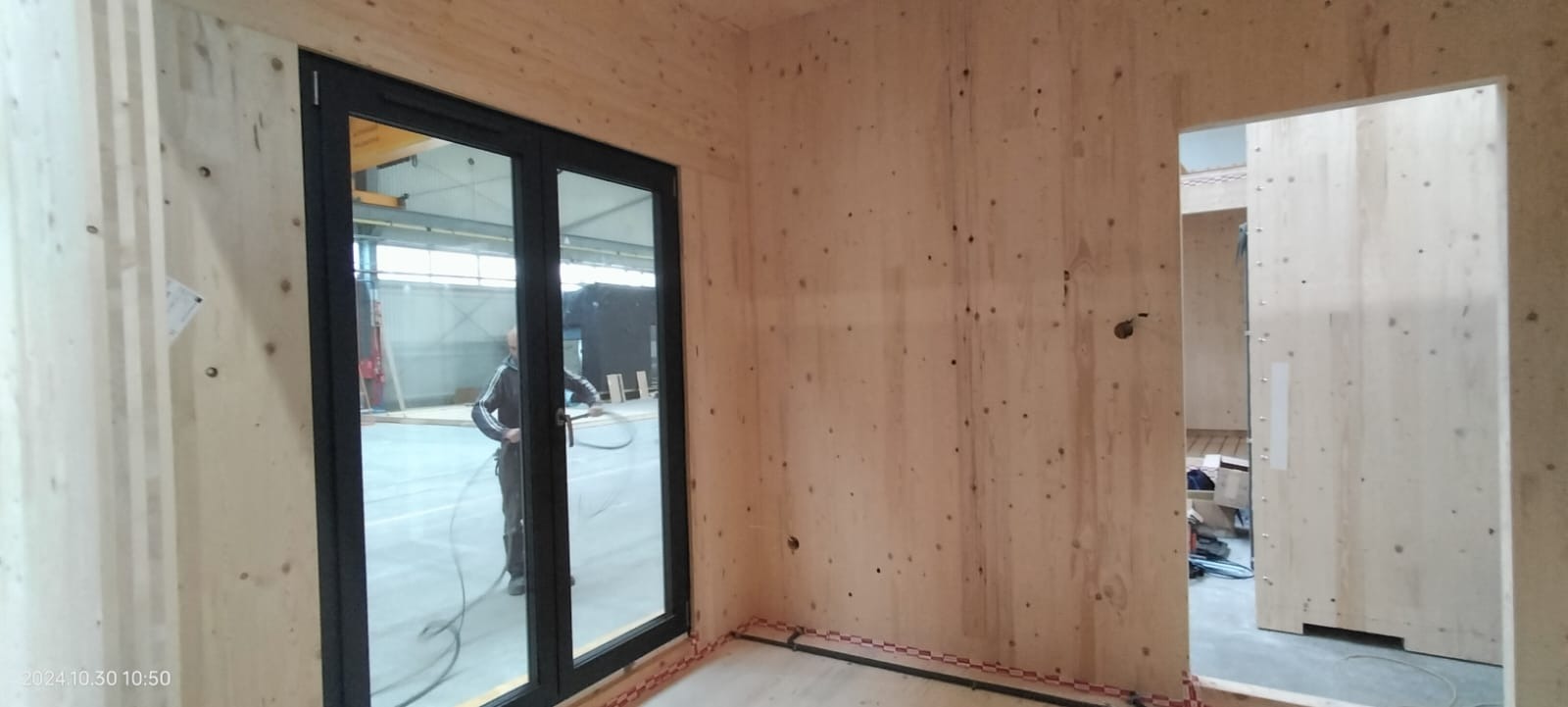 Wnętrze w surowym stanie z drewnianymi ścianami i czarnymi drzwiami balkonowymi; widoczny pracownik z wężem instalacyjnym w tle hali produkcyjnej.