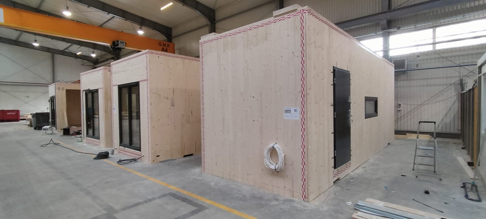 Wnętrze hali produkcyjnej z trzema surowymi modułami domów w trakcie montażu, widoczne okna, drzwi, instalacje elektryczne oraz oznaczenia na ścianach.