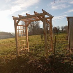 Daniel Sobczyk - Drewniana pergola ogrodowa z kratownicą, wzniesiona na trawniku w słoneczny dzień. W tle zabudowania i drzewa.