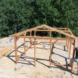 Daniel Sobczyk - Konstrukcja drewnianej altany w trakcie budowy w Rzeszowie. Widoczne elementy szkieletu i przygotowane deski. W tle drzewa i błękitne niebo.