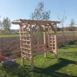 Drewniana pergola z ławką w ogrodzie, obok robot koszący trawę. W tle widoczne pole i drzewa, błękitne niebo. Konstrukcja z jasnego drewna, w stylu rustykalnym.