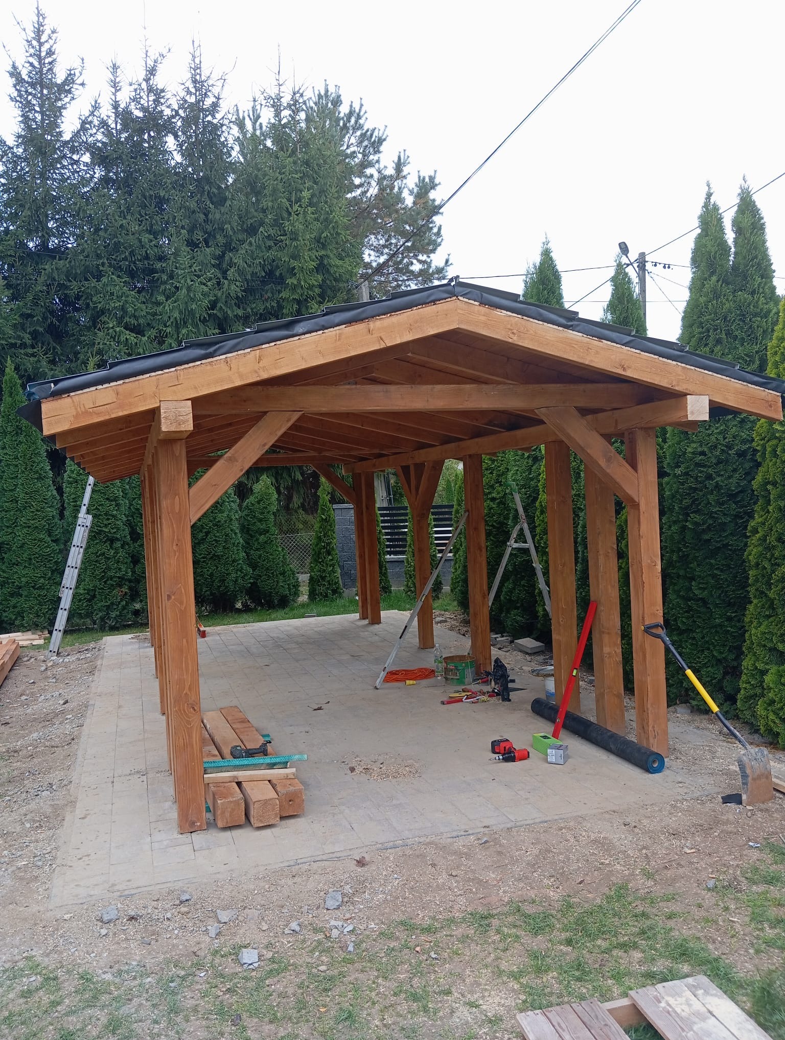 Drewniana altana w trakcie budowy, widoczne narzędzia i materiały budowlane, tło z zielonych drzew iglastych. Częściowo ukończona konstrukcja.