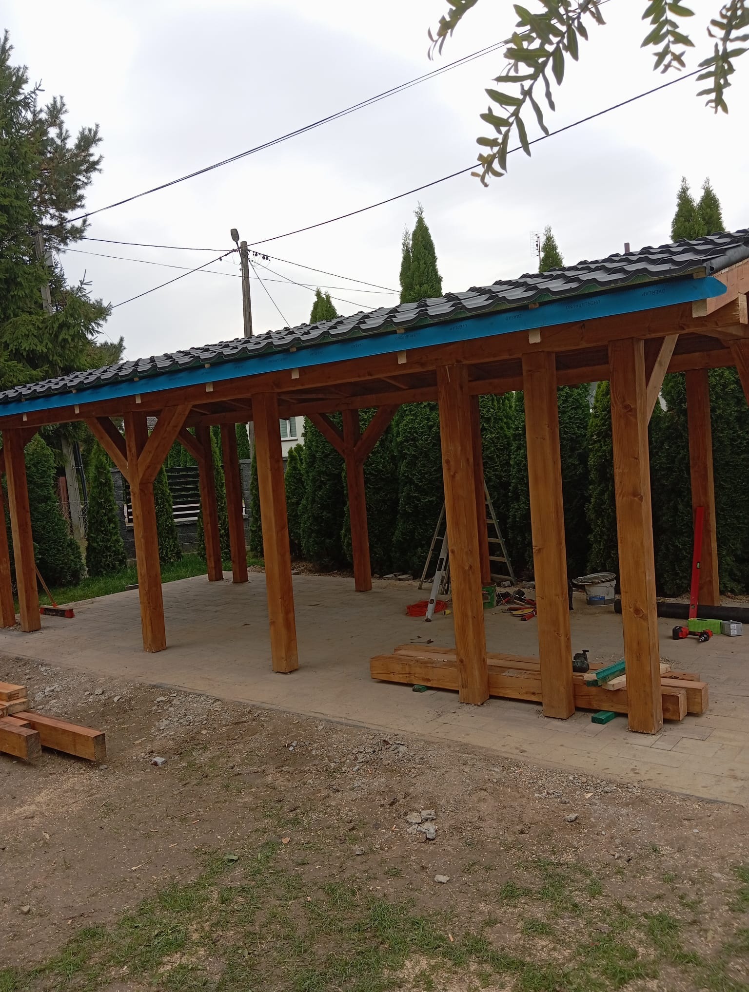 Drewniana konstrukcja w trakcie budowy z widocznym dachem z blachy, narzędziami i otoczeniem zieleni. Widok na ogród i elementy konstrukcyjne.