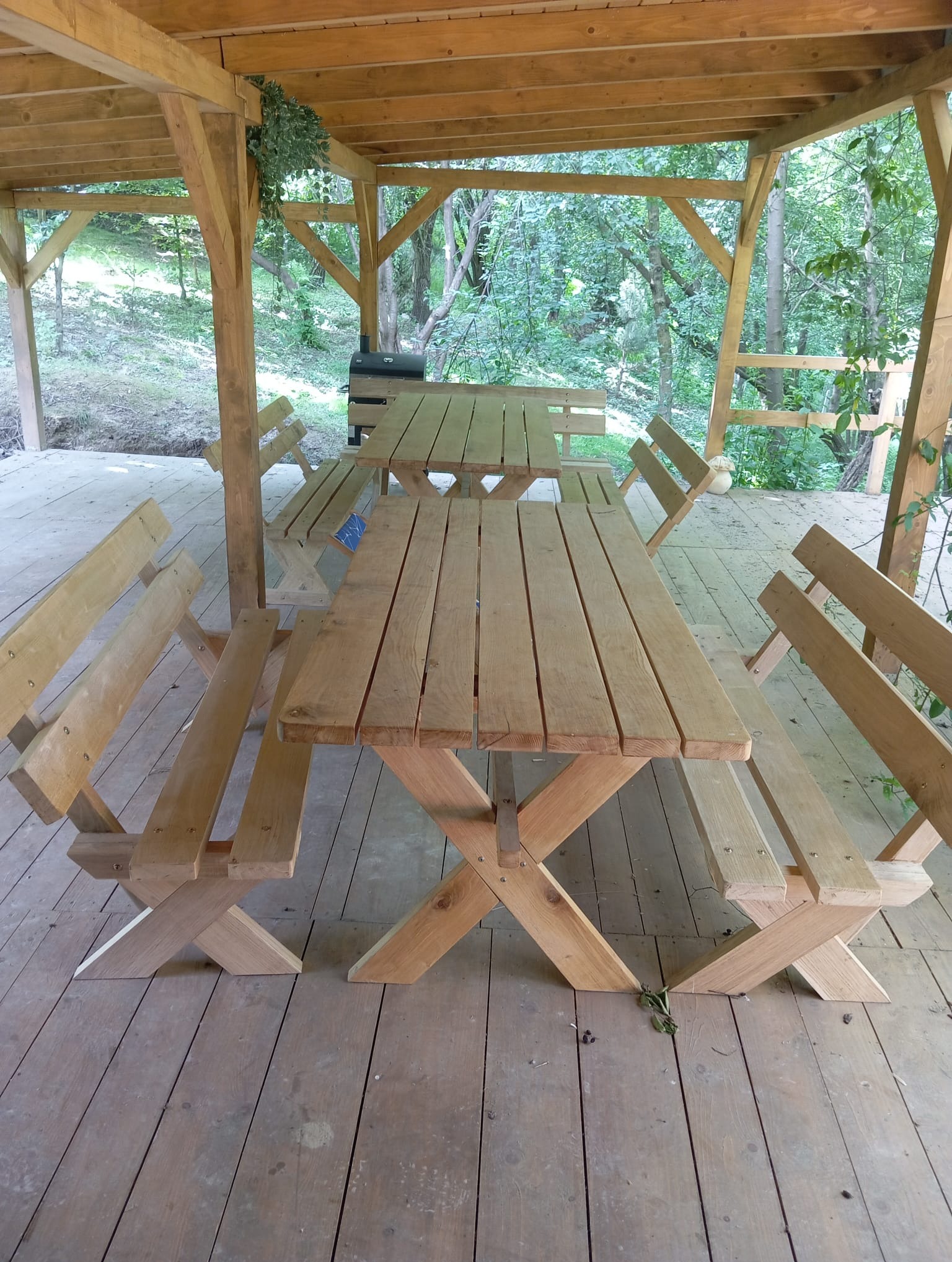 Drewniany stół piknikowy z ławkami pod zadaszeniem, widok na zieleń. Konstrukcja z belek, rustykalny styl, naturalne otoczenie. Detale drewna.