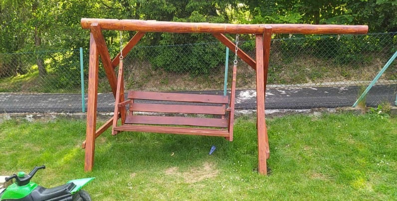 Drewniana huśtawka ogrodowa z surowych bali, zawieszona na łańcuchach, na tle zielonego trawnika i siatki ogrodzeniowej. W tle widoczna droga.