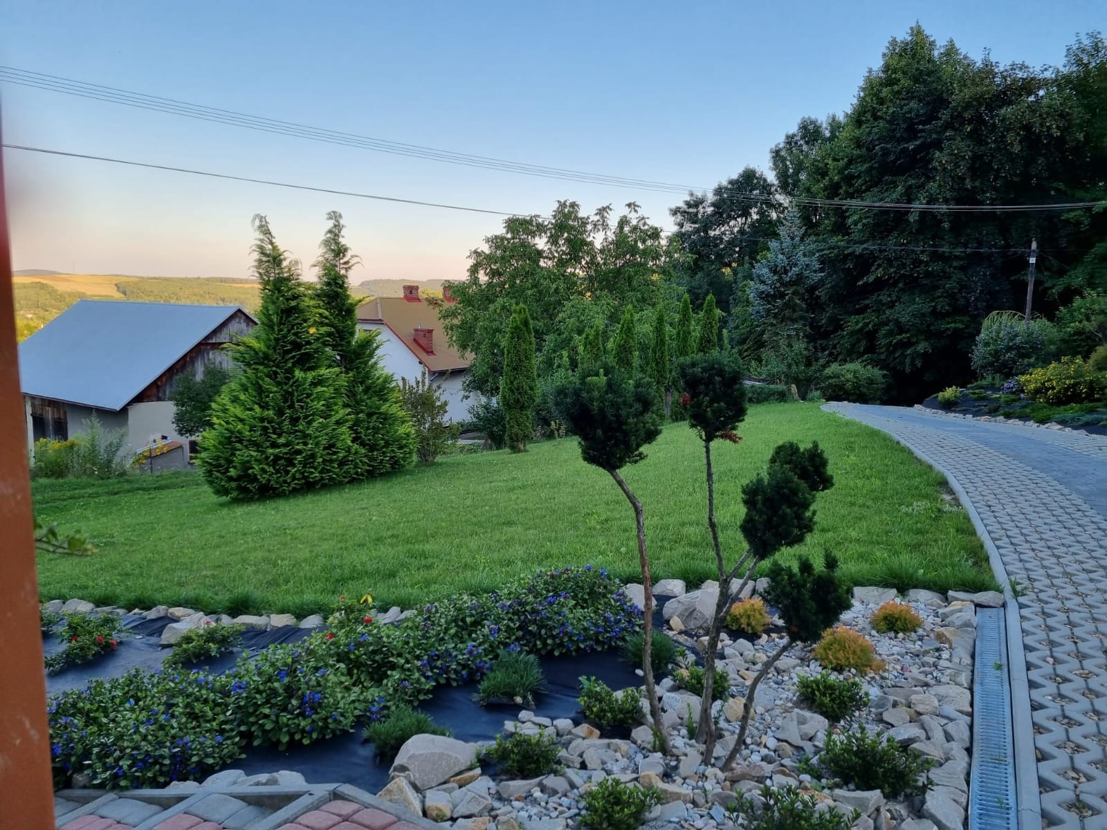 Widok na zadbany ogród z równo przystrzyżonym trawnikiem, ozdobnymi krzewami i ścieżką z kostki brukowej, w tle zabudowania i pagórkowaty krajobraz.