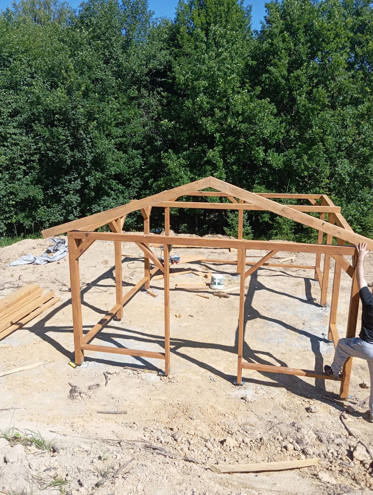 Konstrukcja drewnianej altany w trakcie budowy w Rzeszowie. Widoczne elementy szkieletu i przygotowane deski. W tle drzewa i błękitne niebo.
