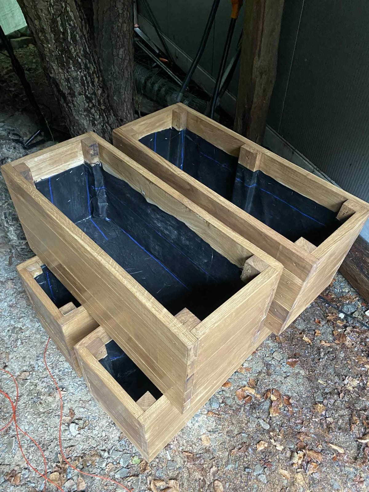 Drewniane, ręcznie robione skrzynie na rośliny z wyściółką wewnątrz, ustawione na zewnątrz, gotowe do sadzenia. Widoczne detale konstrukcyjne i faktura drewna.