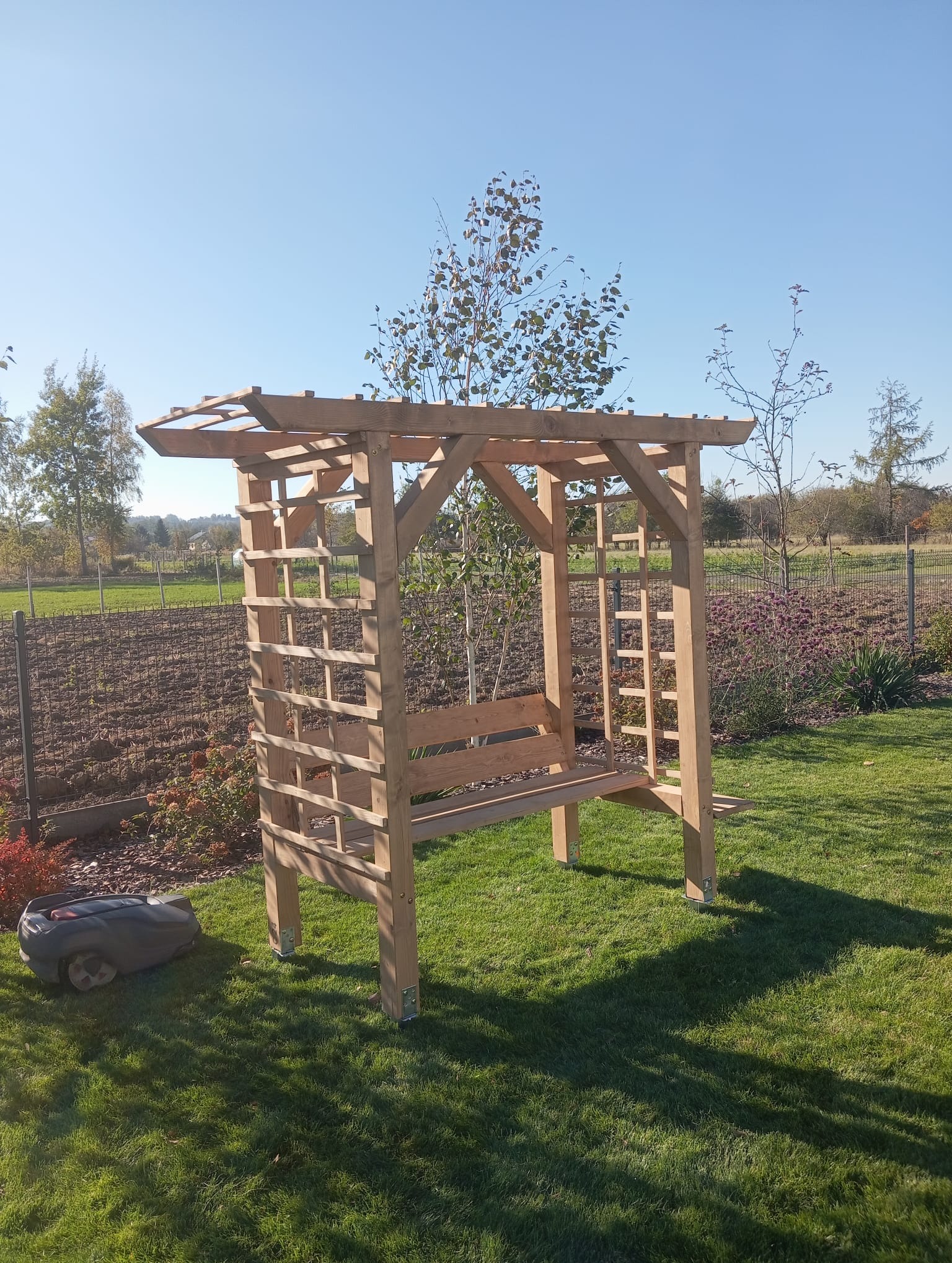 Drewniana pergola z ławką w ogrodzie, obok robot koszący trawę. W tle widoczne pole i drzewa, błękitne niebo. Konstrukcja z jasnego drewna, w stylu rustykalnym.