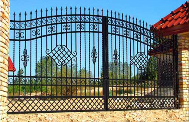Czarna, metalowa brama wjazdowa z ozdobnymi elementami w kształcie rombów i kwiatów, widziana od strony posesji, z fragmentem muru z czerwonej cegły i dachem budynku w tle.
