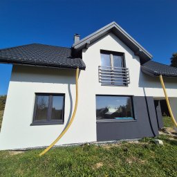 PRYM - Nowoczesny dom z białą elewacją, czarną stolarką okienną i dachem z blachodachówki, z widocznymi żółtymi wężami do ocieplania, stojący na trawiastym terenie.