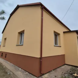 Dwukolorowa elewacja domu jednorodzinnego z brązowym cokołem, beżową górną częścią, brązową rynną i białymi ramami okiennymi, widziana pod kątem w pochmurny dzień.